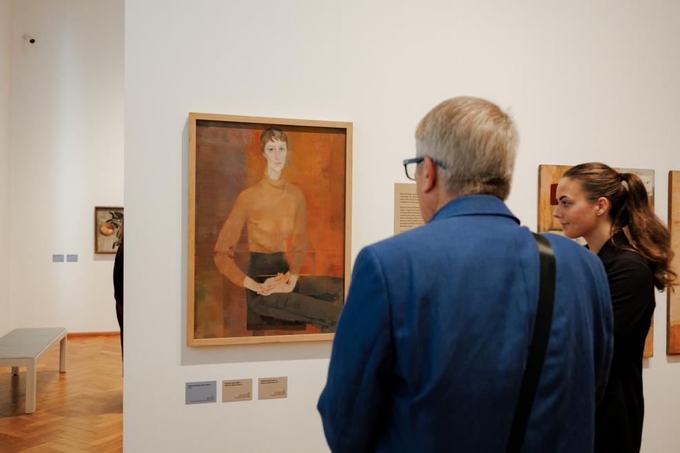 A framed portrait painting in warm orange tones is displayed on a white gallery wall, viewed by two visitors.