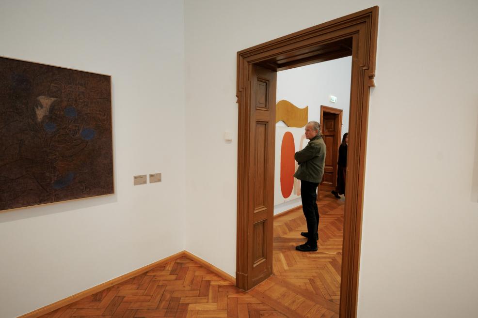 A dark painting hangs on the gallery wall, while through a wooden doorway a man stands beside an abstract work with yellow and orange shapes.