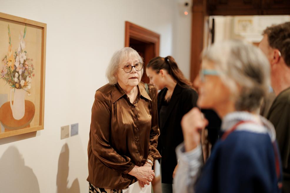 In a museum gallery, a framed floral still life hangs on the wall while an elderly woman wearing glasses stands in the foreground with several visitors.
