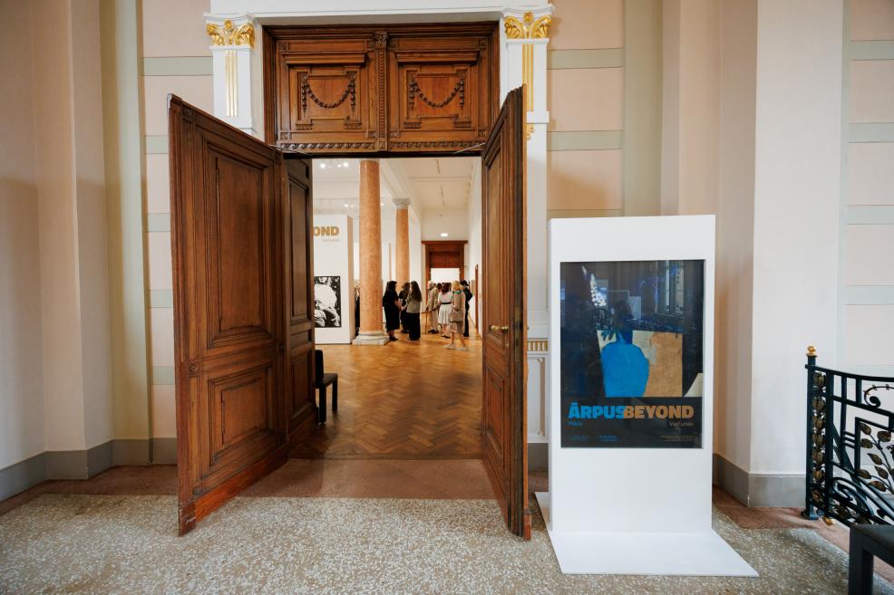 Museum gallery entrance with open wooden double doors, columns and an exhibition poster, with visitors visible in the background.