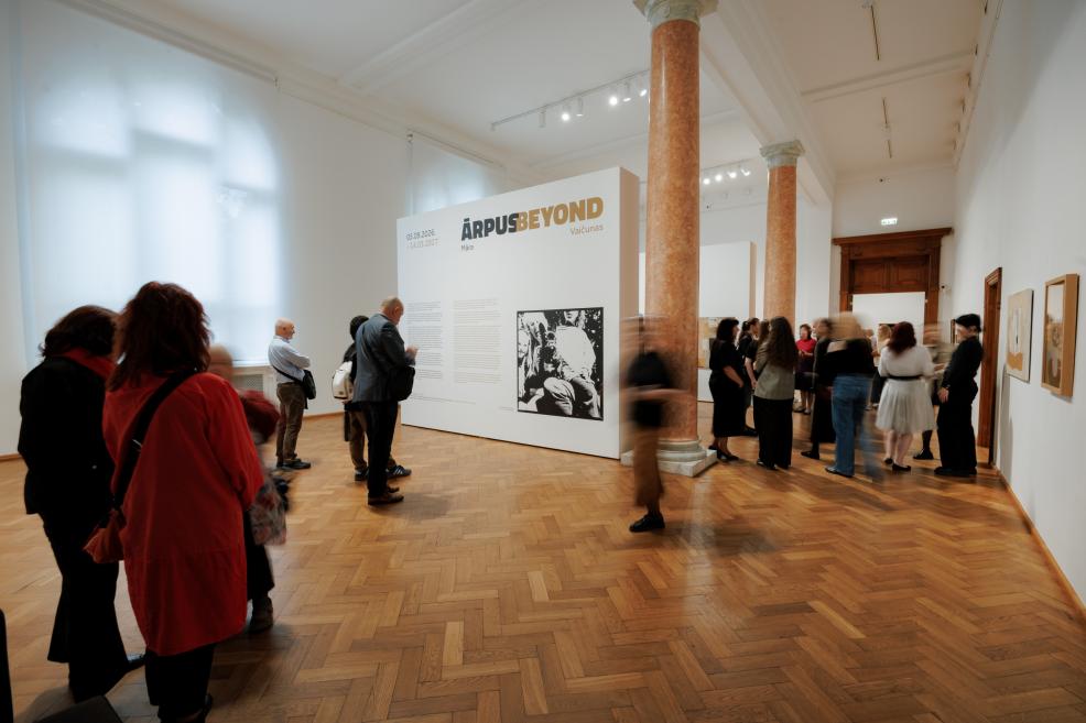 Visitors in a museum gallery view an exhibition beside a large white wall with the text ĀRPUS BEYOND and a black-and-white photograph.