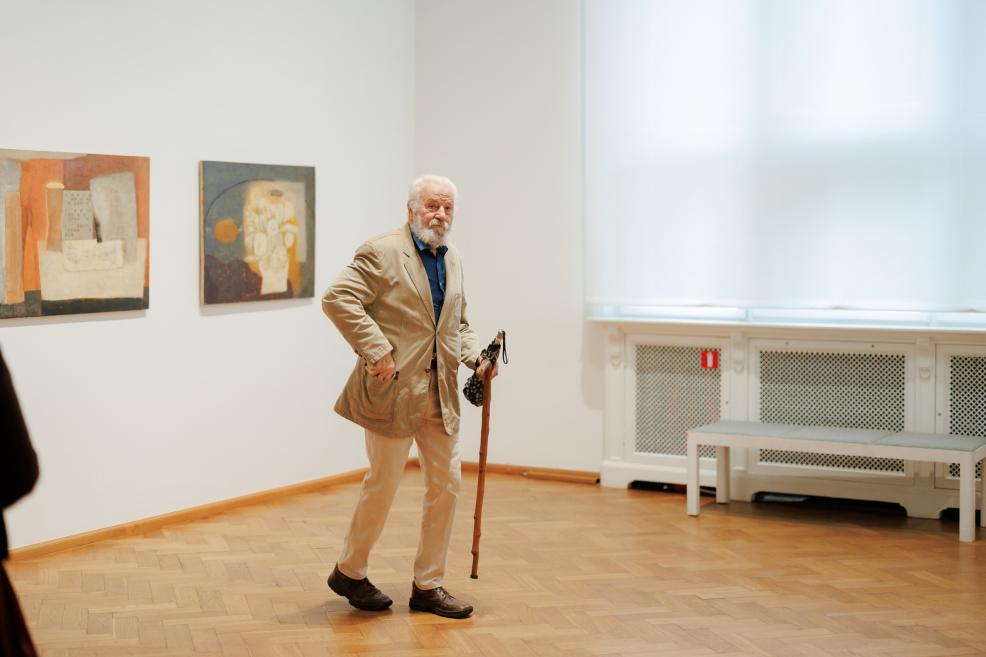 An elderly bearded man with a cane stands in a museum gallery beside two abstract paintings on a white wall.