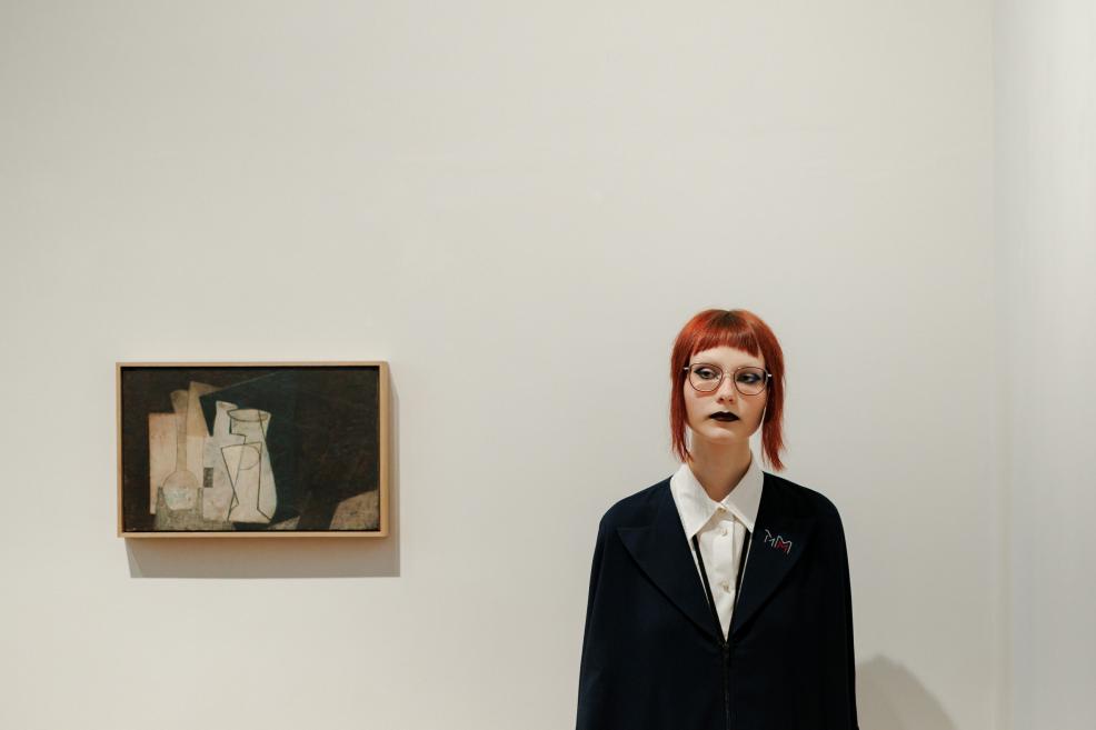 A small abstract painting hangs on a white gallery wall beside a mannequin with red hair, glasses, and a dark suit.
