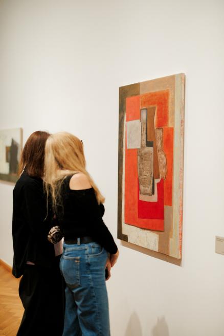 Two women look at an abstract painting with red-orange and gray geometric shapes in a museum gallery.