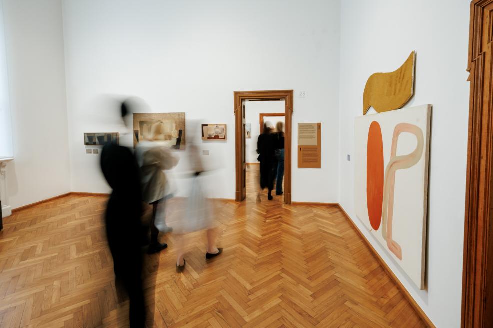 In a museum gallery with parquet flooring and white walls, abstract paintings and installations are displayed while blurred visitors move through the room.
