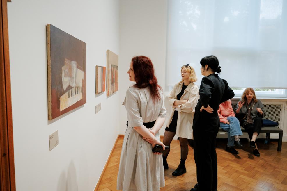 In a museum gallery, several women look at paintings and other small works displayed on a white wall.