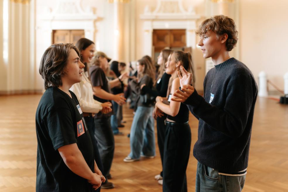 Divi jaunieši sarunājas un gestikulē muzeja zālē, bet fonā vairākas citas personas stāv pāros uz koka grīdas.