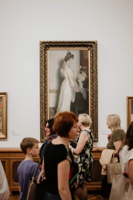 Muzeja zālē pie sienas karājas lielizmēra glezna zeltītā rāmī, kurā attēlota sieviete baltā kleitā, bet priekšplānā stāv apmeklētāji.