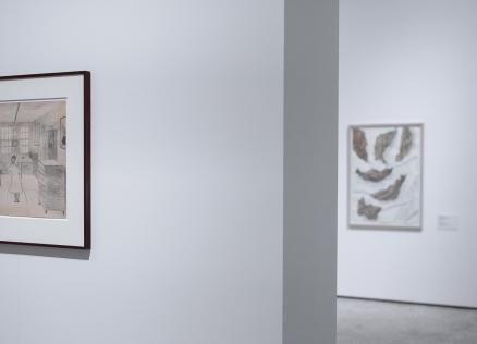 Galerijas zālē pie baltas sienas ierāmēts zīmējums ar interjera ainu, kurā redzama sieviete un suns.