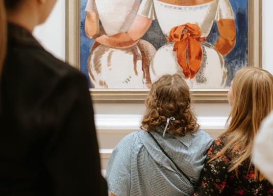 Muzeja zālē vairāki apmeklētāji skatās uz ierāmētu gleznu ar divām sēdošām figūrām baltos tērpos un oranžiem akcentiem.