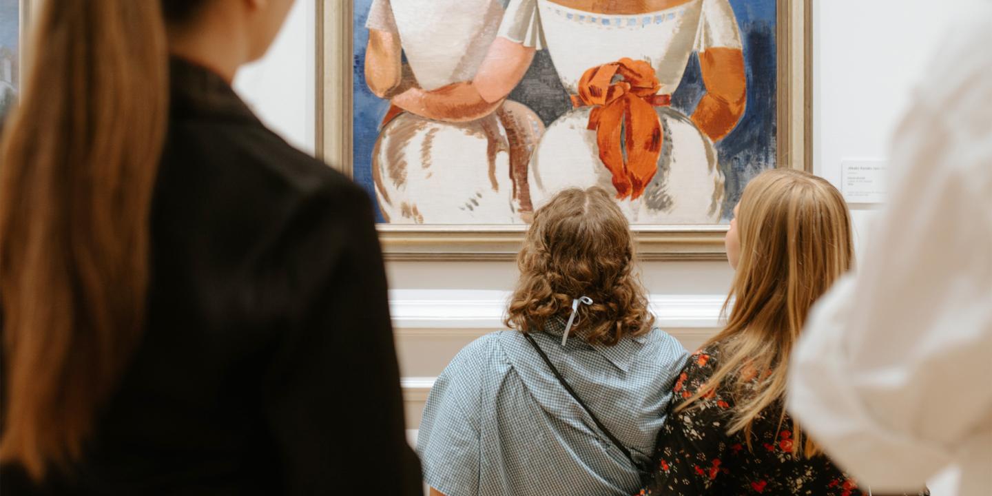 Muzeja zālē vairāki apmeklētāji skatās uz ierāmētu gleznu ar divām sēdošām figūrām baltos tērpos un oranžiem akcentiem.