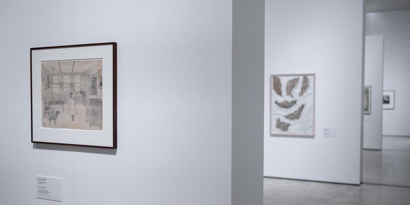 Galerijas zālē pie baltas sienas ierāmēts zīmējums ar interjera ainu, kurā redzama sieviete un suns.