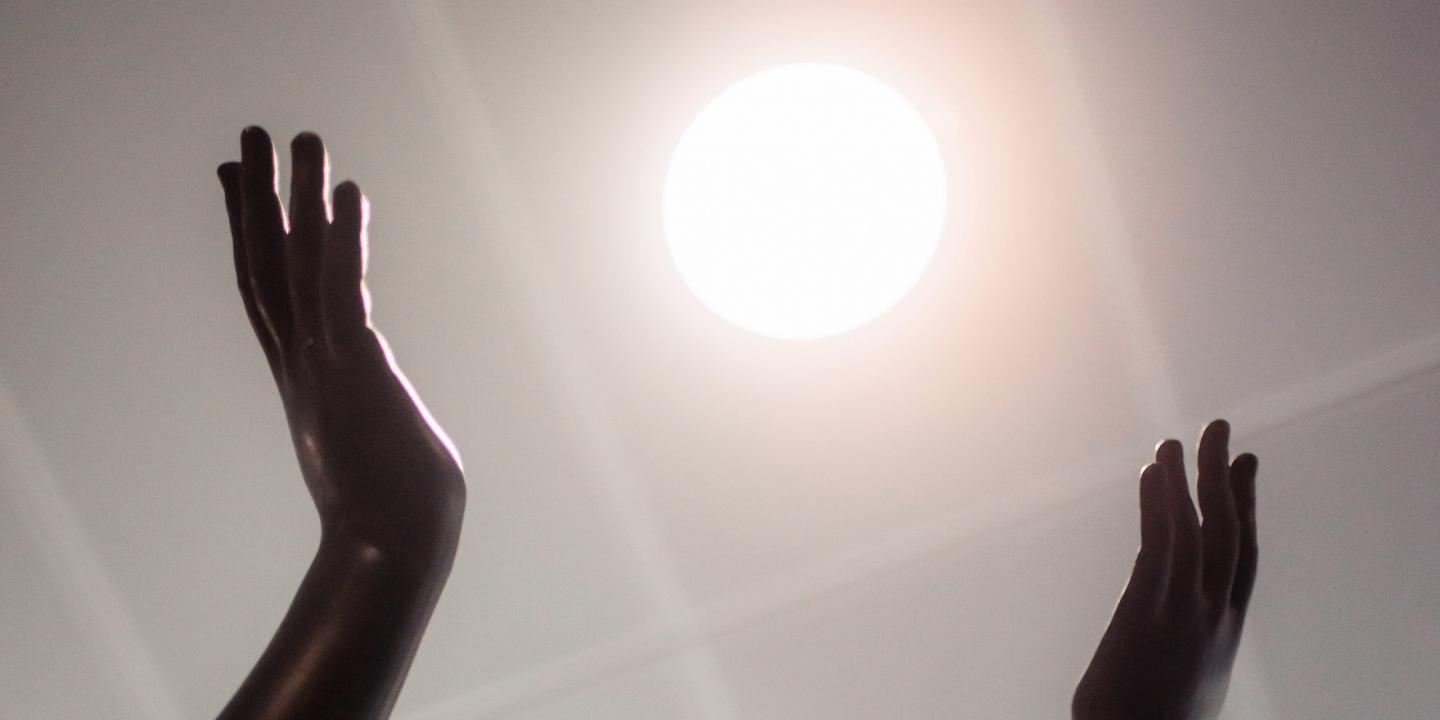 Tumšas skulptūras rokas paceltas pret spožu gaismas apli, kas atgādina sauli.