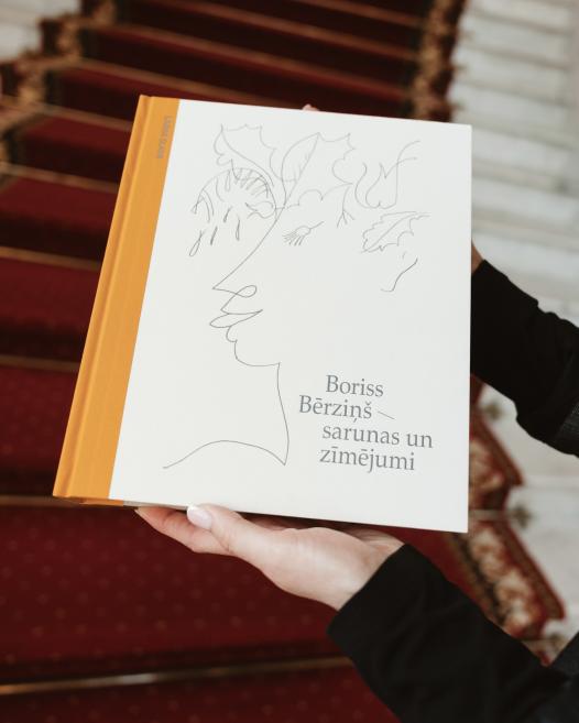 Rokās turēta grāmata “Boriss Bērziņš — sarunas un zīmējumi” uz muzeja kāpņu sarkanā paklāja fona.