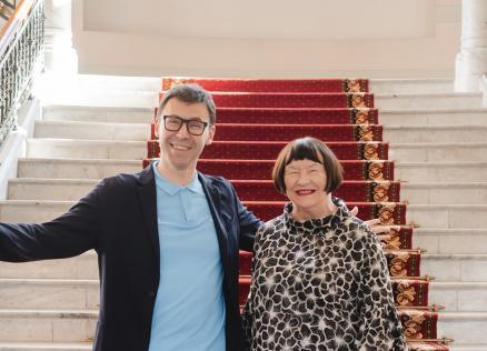 Divi cilvēki stāv uz muzeja galvenajām kāpnēm ar sarkanu paklāju.