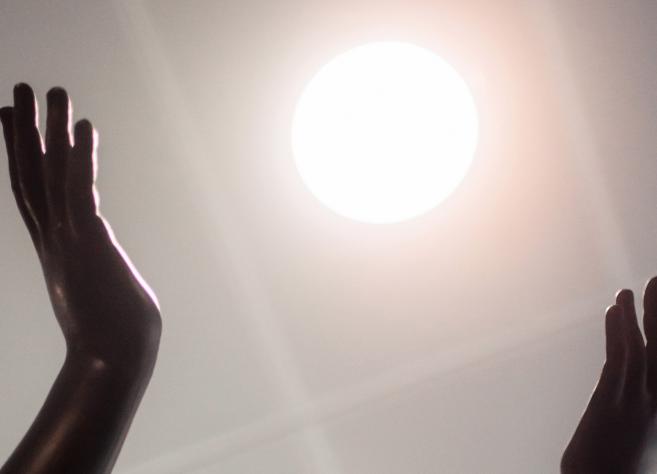 Tumšas skulptūras rokas paceltas pret spožu gaismas apli, kas atgādina sauli.