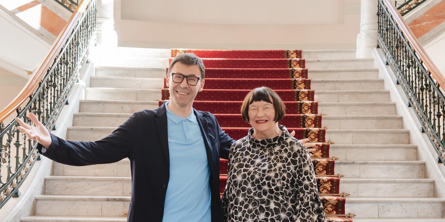 Divi cilvēki stāv uz muzeja galvenajām kāpnēm ar sarkanu paklāju.