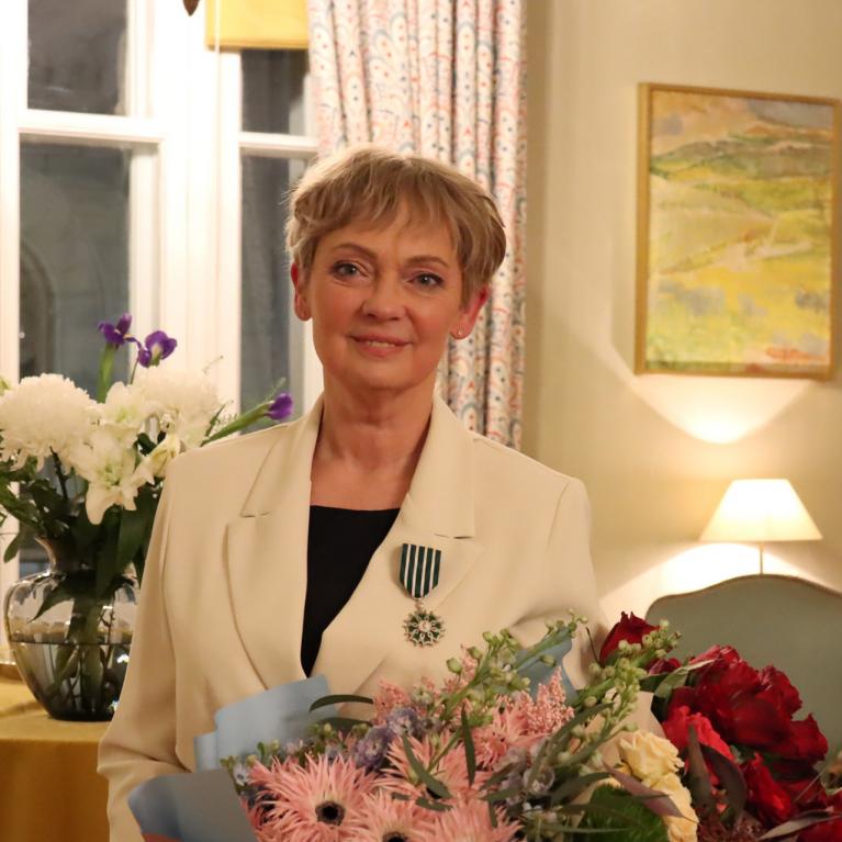 A woman wearing a light-coloured jacket with an order insignia and holding a bouquet stands in a classical interior with candles and flowers.