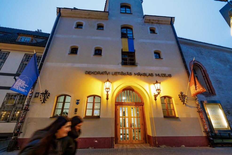 Dekoratīvās mākslas un dizaina muzeja ieejas fasāde vakara apgaismojumā. Vēsturiska ēka ar arkveida durvīm, izgaismotiem sienas lukturiem un karogiem pie ieejas, priekšplānā redzami garāmgājēji kustībā.
