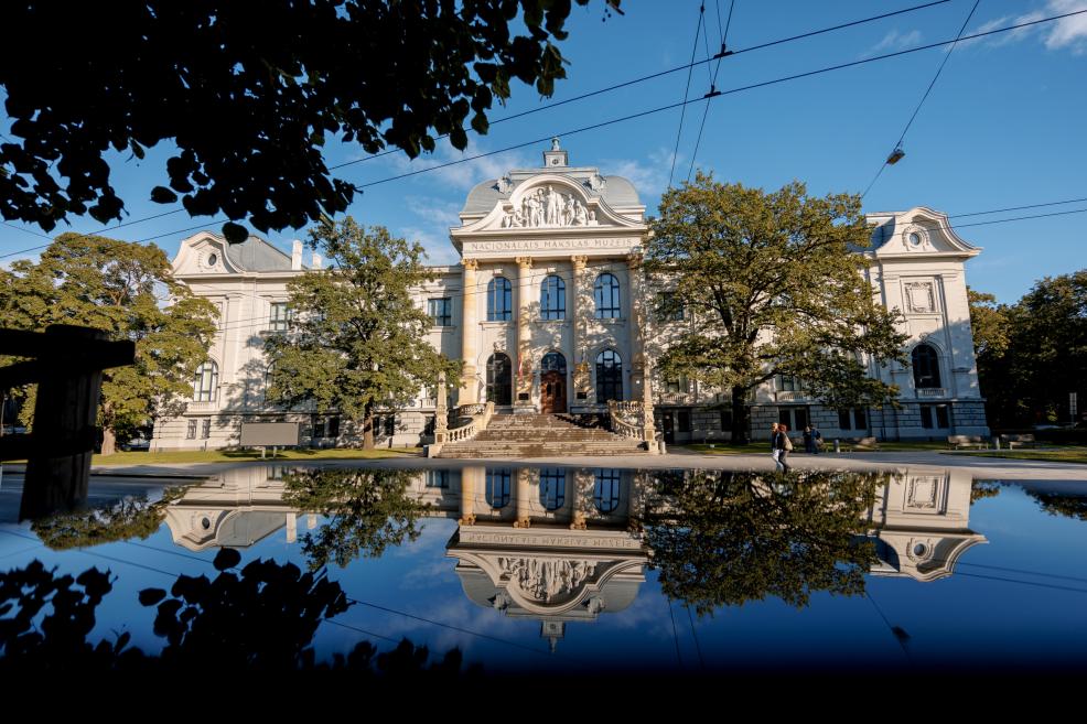 Latvijas Nacionālā mākslas muzeja galvenā ēka saulainā dienā, skatā no parka puses. Vēsturiskā neoklasicisma fasāde ar centrālo portiku un skulpturālu frontonu atspoguļojas ūdens peļķē priekšplānā, apkārt redzami koki un gājēji.