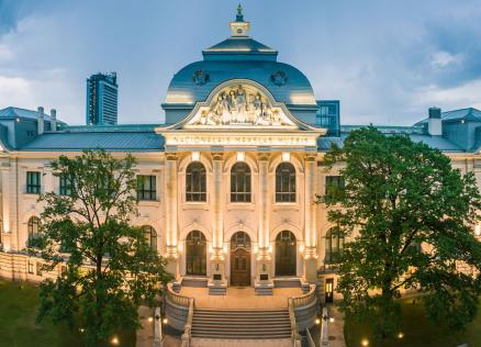 Latvijas Nacionālā mākslas muzeja galvenās ēkas fasāde vakara apgaismojumā. Vēsturiska neoklasicisma stila ēka ar centrālo kupolu, skulpturālu frontonu un simetrisku arhitektūru, ko ieskauj zaļi koki un pilsētas apbūve.