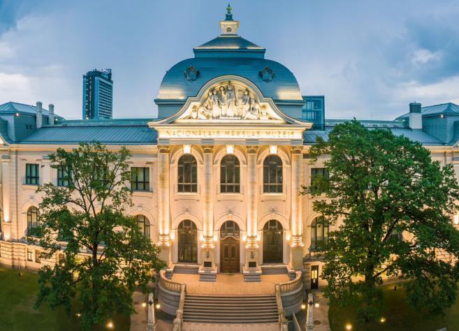 Latvijas Nacionālā mākslas muzeja galvenās ēkas fasāde vakara apgaismojumā. Vēsturiska neoklasicisma stila ēka ar centrālo kupolu, skulpturālu frontonu un simetrisku arhitektūru, ko ieskauj zaļi koki un pilsētas apbūve.