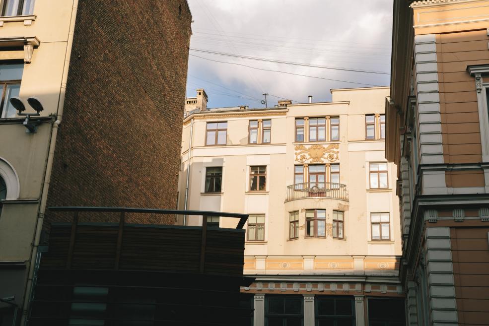 Vēsturisku pilsētas ēku iekšpagalma skats ar daudzstāvu namu fasādēm. Centrā redzama gaiša ēka ar dekoratīviem elementiem un balkonu, apkārt – šaurā telpā izvietotas citas ēkas, virs tām – debesu fragments un elektrības vadi.