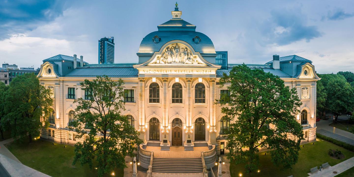 Latvijas Nacionālā mākslas muzeja galvenās ēkas fasāde vakara apgaismojumā. Vēsturiska neoklasicisma stila ēka ar centrālo kupolu, skulpturālu frontonu un simetrisku arhitektūru, ko ieskauj zaļi koki un pilsētas apbūve.