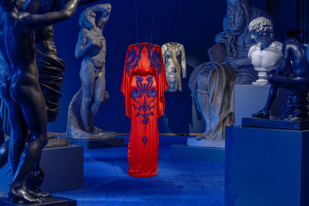 Zilā izstāžu zālē uz pjedestāliem un pie sienām izvietotas skulptūras un divi dekoratīvi tērpi, kuru vidū karājas spilgti sarkans kleitas tērps ar ziliem ornamentiem.