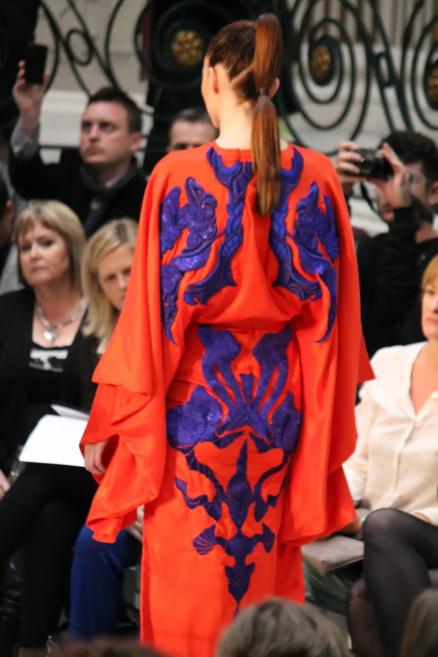 Modele uz skrejceļa oranžā tērpā ar tumši violetiem ornamentiem, skatīta no mugurpuses skatītāju priekšā.