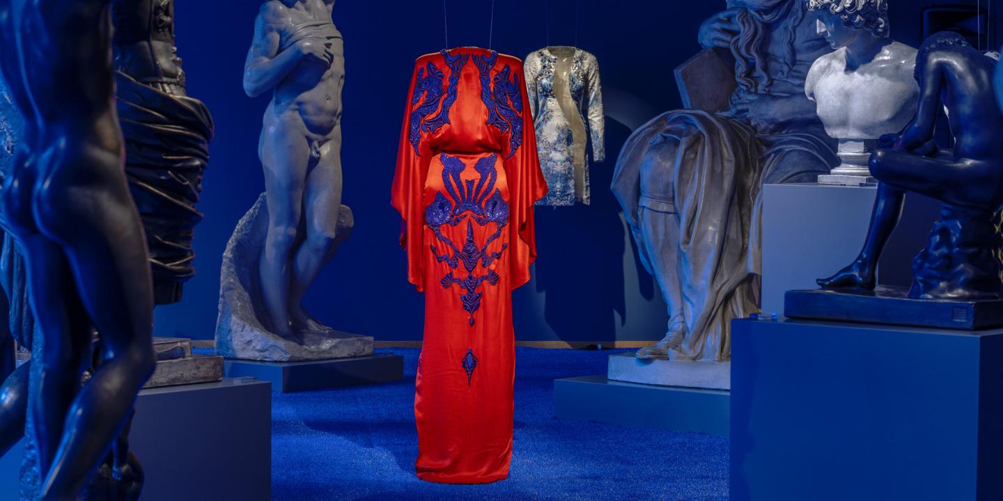 Izstāžu zālē uz zila fona starp skulptūrām uz pakaramā redzama koši sarkana gara kleita ar tumši ziliem ornamentiem.