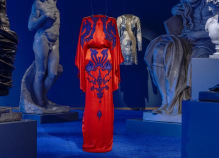 Izstāžu zālē uz zila fona starp skulptūrām uz pakaramā redzama koši sarkana gara kleita ar tumši ziliem ornamentiem.