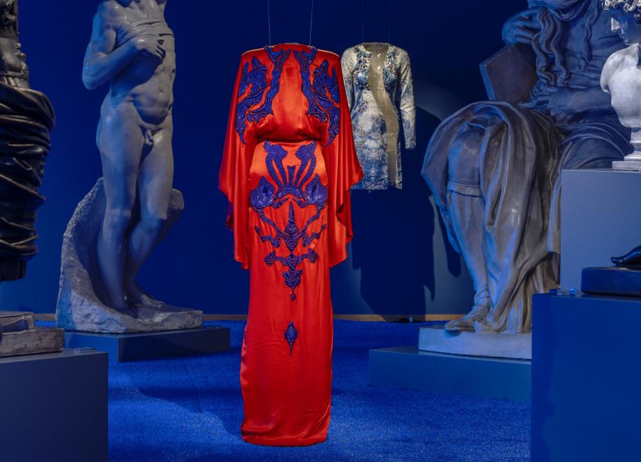 Izstāžu zālē uz zila fona starp skulptūrām uz pakaramā redzama koši sarkana gara kleita ar tumši ziliem ornamentiem.