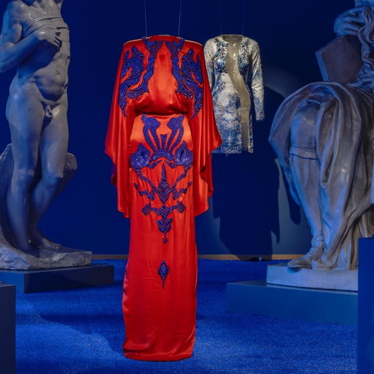 Izstāžu zālē uz zila fona starp skulptūrām uz pakaramā redzama koši sarkana gara kleita ar tumši ziliem ornamentiem.