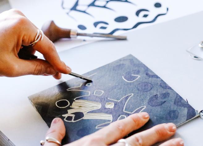 Rokas ar instrumentu skrāpē attēlu uz tumšas virsmas, veidojot baltas līnijas un formas, apkārt redzami grafikas rīki un izgriezumi.