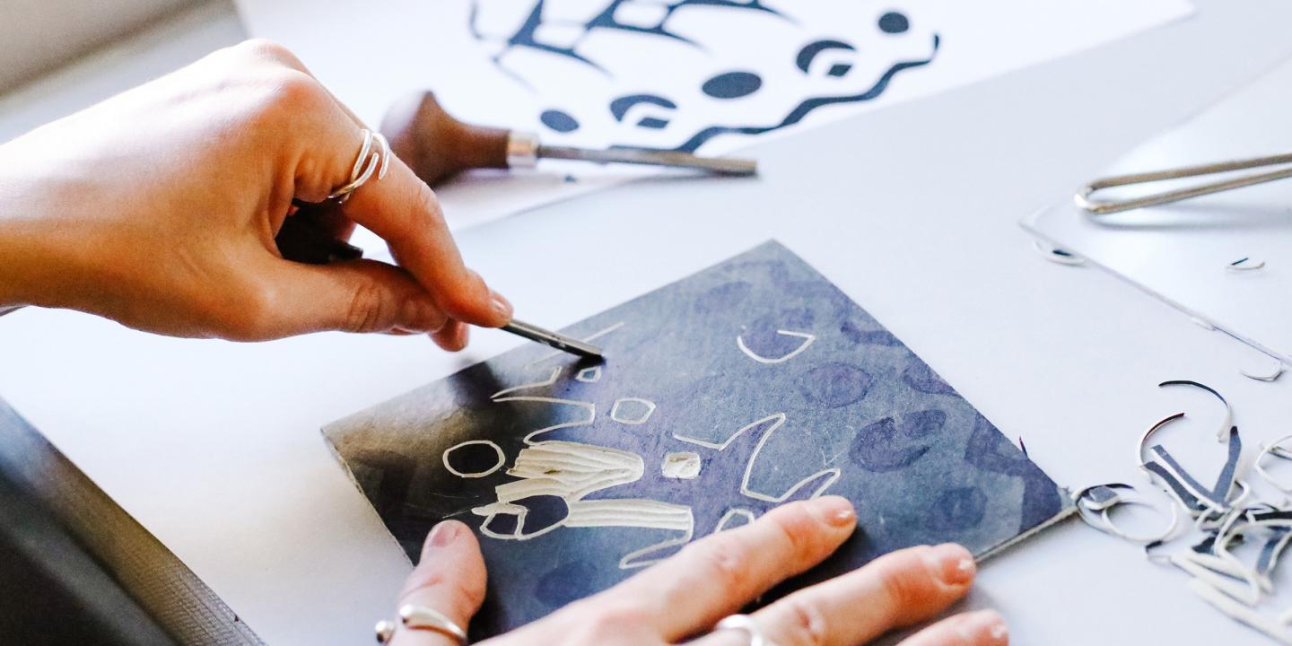 Rokas ar instrumentu skrāpē attēlu uz tumšas virsmas, veidojot baltas līnijas un formas, apkārt redzami grafikas rīki un izgriezumi.