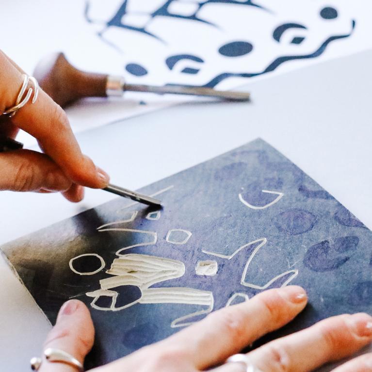 Rokas ar instrumentu skrāpē attēlu uz tumšas virsmas, veidojot baltas līnijas un formas, apkārt redzami grafikas rīki un izgriezumi.