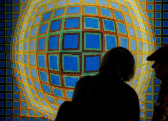 Divi apmeklētāji siluetā aplūko ģeometrisku, krāsainu gleznu ar režģveida sfērisku formu uz muzeja sienas.