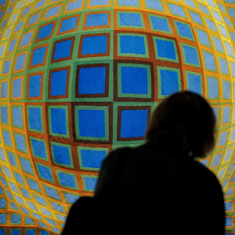 Divi apmeklētāji siluetā aplūko ģeometrisku, krāsainu gleznu ar režģveida sfērisku formu uz muzeja sienas.