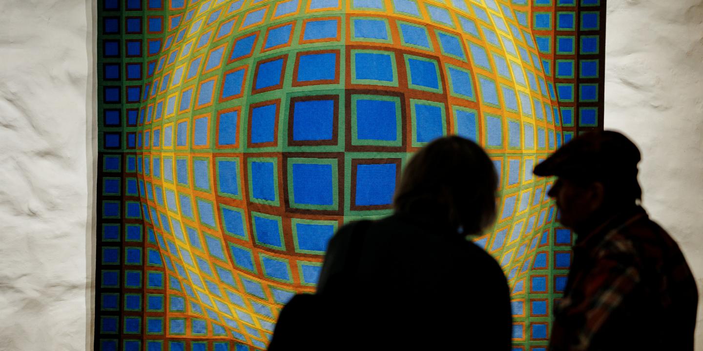 Divi apmeklētāji siluetā aplūko ģeometrisku, krāsainu gleznu ar režģveida sfērisku formu uz muzeja sienas.