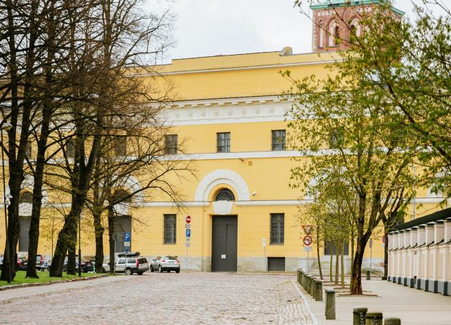 Latvijas Nacionālā mākslas muzeja dzeltenā ēka ar arku veida ieeju, redzama starp kokiem bruģētas ielas malā.