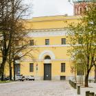 Latvijas Nacionālā mākslas muzeja dzeltenā ēka ar arku veida ieeju, redzama starp kokiem bruģētas ielas malā.