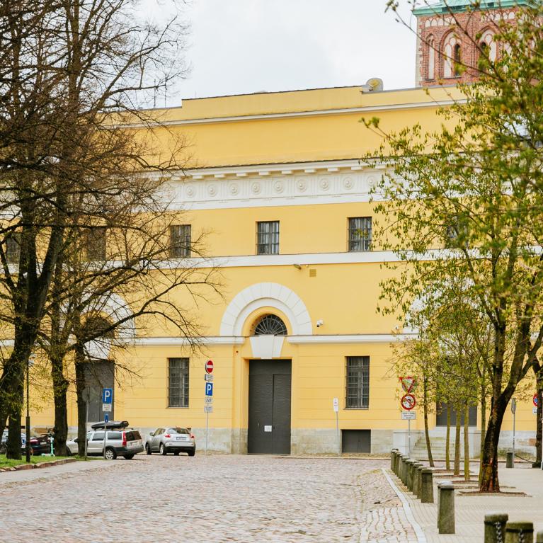 Latvijas Nacionālā mākslas muzeja dzeltenā ēka ar arku veida ieeju, redzama starp kokiem bruģētas ielas malā.
