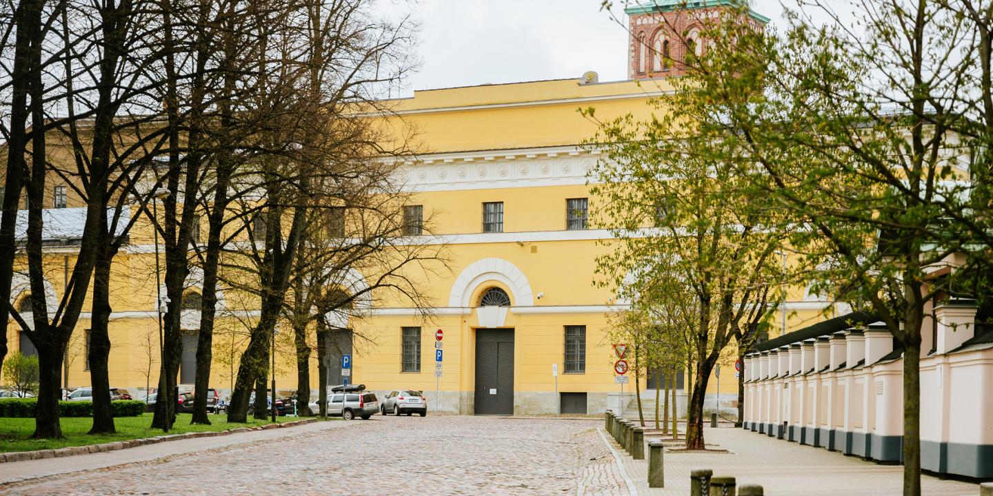 Latvijas Nacionālā mākslas muzeja dzeltenā ēka ar arku veida ieeju, redzama starp kokiem bruģētas ielas malā.