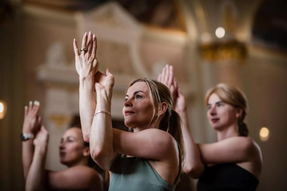 Three women practice yoga in a museum hall, performing an Eagle Pose arm variation with focused expressions and hands raised before their faces.