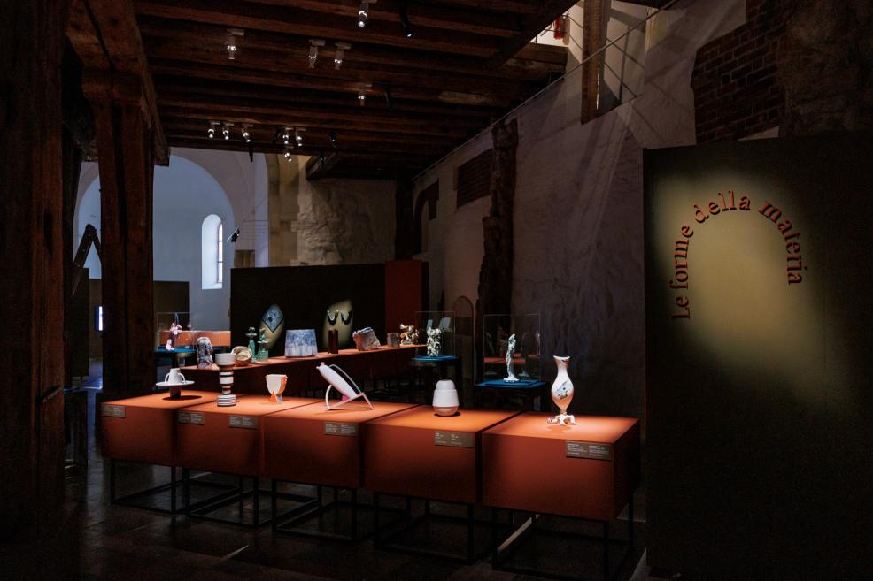 Various ceramic and design objects are displayed on orange pedestals and in vitrines in a museum exhibition hall, with the words Le forme della materia on a dark panel.