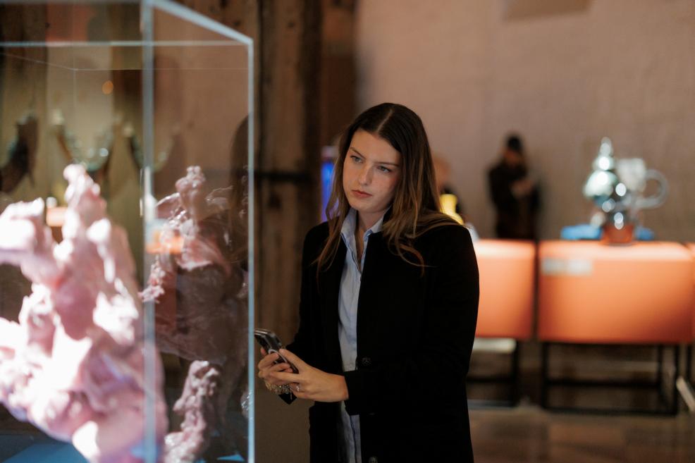 A young woman in a black blazer looks at a pink sculptural object displayed in a glass case at a museum exhibition.