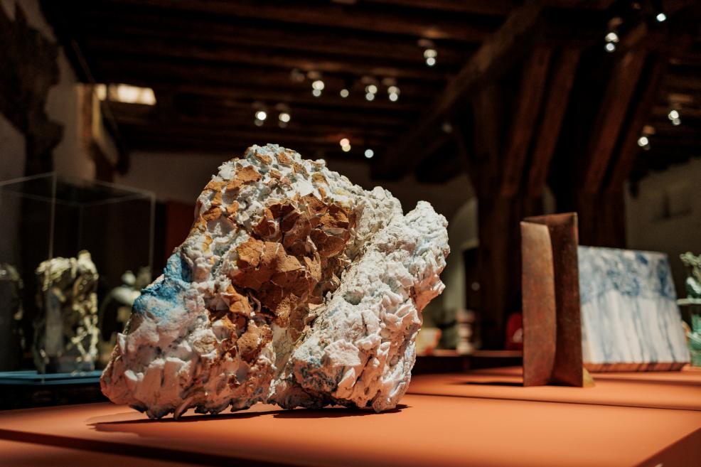 A large rough-surfaced sculpture in white, blue and brown tones stands on an orange pedestal in a museum exhibition hall.