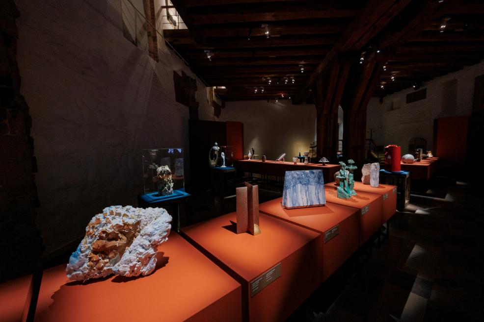 Museum exhibition hall with various sculptures and objects displayed on orange pedestals beneath dark wooden ceiling beams.