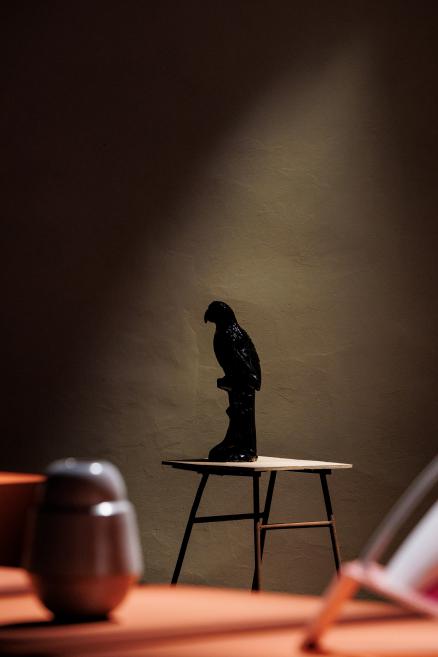 A dark bird sculpture on a small table against an illuminated wall, with blurred seats in the foreground.